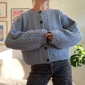 Könnte beinhalten: Hellblauer Zopfstrick-Cardigan mit dunkelbraunen Knöpfen. Der Pullover hat einen runden Ausschnitt und lange Ärmel. Die Person trägt Jeans mit Leopardenmuster. Der Hintergrund umfasst gerahmte Kunstwerke und eine Zimmerpflanze.