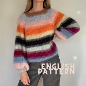 Könnte beinhalten: Ein langärmeliger, flauschiger Strickpullover mit horizontalen Streifen in den Farben Braun, Orange, Gelb, Schwarz, Hellblau und Magenta. Der Pullover hat einen Rundhalsausschnitt und gerippte Bündchen. Der Text "ENGLISH PATTERN" ist unten rechts zu sehen.