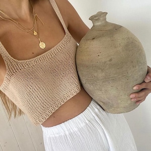 Op de afbeelding: Een vrouw draagt een beige gebreid crop top en een witte broek en houdt een grote, ronde, bruine kleipot vast.