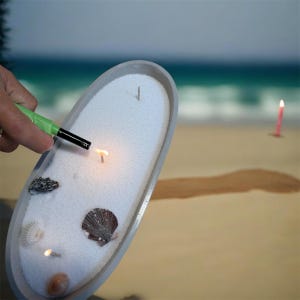 May include: An oval gray tray filled with white sand, featuring several seashells and multiple candle wicks. A green lighter is being used to ignite one of the wicks, creating a small flame. The tray is held against a blurred beach background.