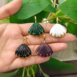 May include: Four fan-shaped pendants in dark green, white, black, and purple, each featuring vertical carved ridges and a gold-toned metal setting with a top bail. The pendants are arranged in a cluster, showcasing their smooth, polished surfaces.