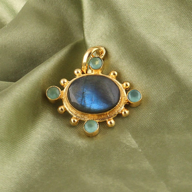 May include: A gold-colored pendant with a large, oval, iridescent blue-gray stone at its center. The pendant is adorned with smaller, round, light blue-green stones and decorative gold accents. The pendant is set against a green fabric background.