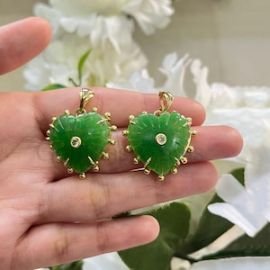 May include: Two heart-shaped green jade pendants with gold-colored accents. Each pendant features a central diamond and a gold bail. The edges are adorned with small gold beads. The pendants are displayed against a soft, blurred background.