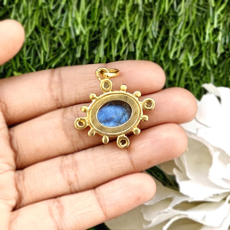 May include: A gold-colored pendant with an oval, blue gemstone center. The pendant has a decorative sunburst design around the gemstone. The pendant is held in a person's hand.