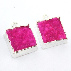 May include: Two square-shaped, silver-toned pendants with a vibrant pink, crystalline center. The edges of the pink crystal are uneven, and the pendants have a small loop at the top for hanging. The pendants are set against a white background.