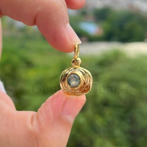 以下が含まれることがあります： 中央に淡い色の宝石をあしらった、円形デザインのゴールドカラーのペンダント。ぼやけた緑色の背景を背景に、指でペンダントを保持しています。「Gun Gems」の文字が見えます。