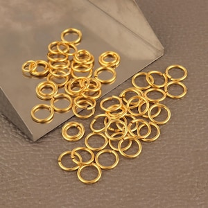 May include: A large quantity of gold jump rings, a type of jewelry finding, are scattered on a gray surface. The rings are small and circular, perfect for connecting jewelry components.