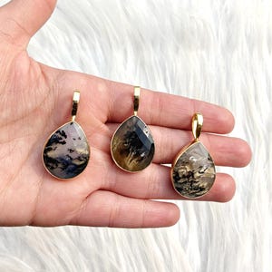 May include: Three teardrop-shaped pendants with gold-colored settings. The pendants feature unique patterns in shades of black, gray, and brown. The pendants are displayed against a white, fluffy background.