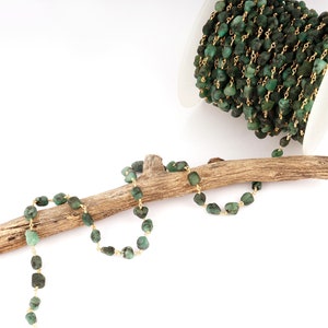 Peut inclure: Cha&icirc;ne de pierres pr&eacute;cieuses vert &eacute;meraude avec fil dor&eacute;, pr&eacute;sent&eacute;e sur un morceau de bois flott&eacute;. Une bobine de la cha&icirc;ne est visible en arri&egrave;re-plan. La cha&icirc;ne est compos&eacute;e de petites pierres de couleur &eacute;meraude irr&eacute;guli&egrave;res.