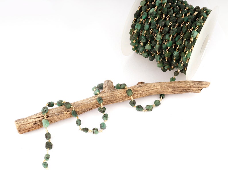Peut inclure: Cha&icirc;ne de pierres pr&eacute;cieuses vert &eacute;meraude avec fil dor&eacute;, pr&eacute;sent&eacute;e sur un morceau de bois flott&eacute;. Une bobine de la cha&icirc;ne est visible en arri&egrave;re-plan. La cha&icirc;ne est compos&eacute;e de petites pierres de couleur &eacute;meraude irr&eacute;guli&egrave;res.