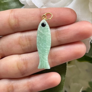 May include: A light green fish-shaped pendant with a gold-colored loop. The pendant has a black eye and a textured surface. The fish pendant is held in a hand.