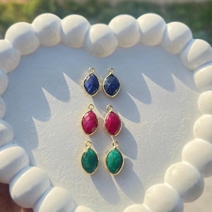 May include: Six gemstone pendants in blue, red, and green, each with a gold-colored bezel. The teardrop-shaped stones are faceted, catching the light. The pendants are displayed on a white, scalloped surface.