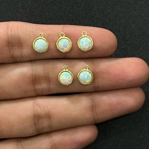 Könnte beinhalten: Sechs kleine, runde, goldfarbene Anhänger mit irisierenden, opalartigen Zentren. Jeder Anhänger hat oben eine kleine Schlaufe zum Befestigen an Schmuck. Die Anhänger werden auf neutralem Hintergrund präsentiert.