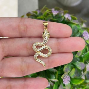 以下が含まれることがあります： 白エナメルとクリアクリスタルアクセントが施されたゴールドカラーのヘビペンダント。ヘビの頭は黒と白で、体はコイル状になっています。ペンダントはネックレスとして着用するように設計されています。