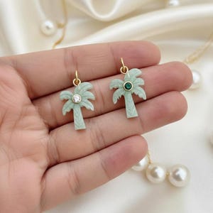 May include: Two light green palm tree charms with gold-colored loops. One charm has a clear crystal, the other a green gem. The charms are displayed on a white surface with a pearl necklace.