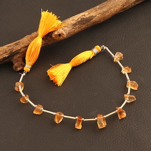 Puede incluir: Un collar con piedras preciosas de citrino en bruto de color ámbar ensartadas en un alambre plateado. También hay dos borlas de color amarillo brillante. El collar y las borlas se exhiben sobre una superficie marrón oscuro con un trozo de madera.