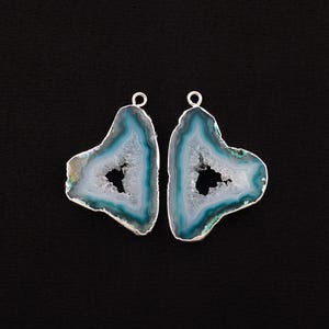 Puede incluir: Dos colgantes de ágata pulida en forma de corazón con un borde plateado. Los colgantes presentan un centro blanco, rodeado de bandas azules y turquesas, sobre un fondo gris oscuro.