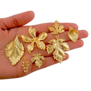 May include: A collection of gold-colored leaf and flower charms. The charms vary in size and design, including a large flower, several different leaf shapes, and a small leaf with pearl-like accents. The charms are held in a hand.