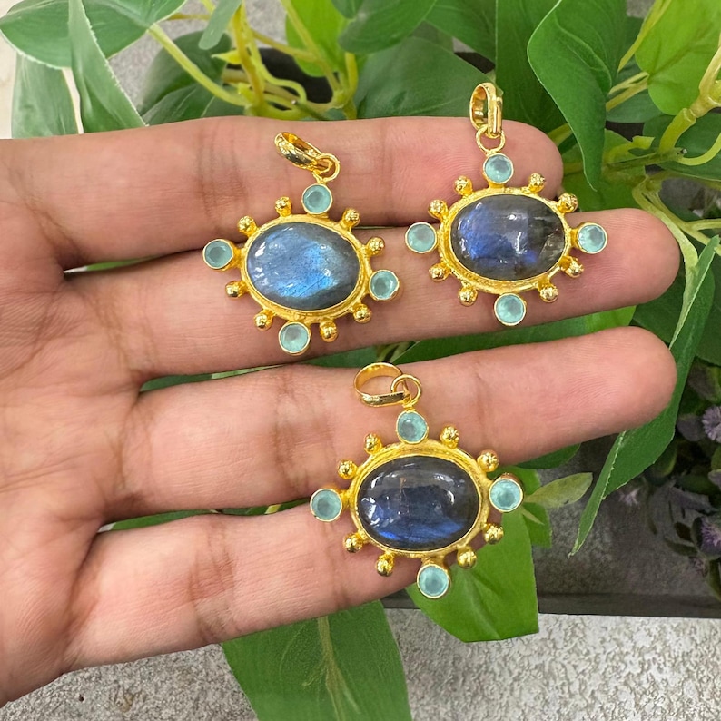 May include: Three gold-colored pendants with oval, iridescent blue gemstones. Each pendant is surrounded by small, light blue gemstones and a sunburst design. The pendants are displayed against a background of green leaves.