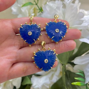 May include: Three heart-shaped pendants with a vibrant blue hue. Each pendant features a gold-colored bail and a central, clear stone. The edges are adorned with small, gold-colored accents. The pendants are displayed against a backdrop of white flowers.