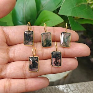 May include: Five rectangular pendants featuring moss agate gemstones with dark green and black dendritic inclusions. Each gemstone is set in a gold-toned metal bezel with a matching gold-toned jump ring attached to the top for hanging purposes.