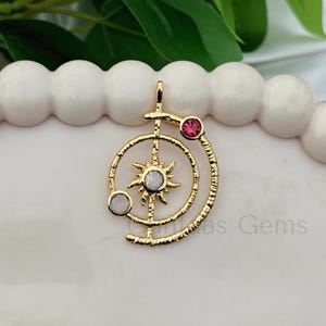 Pode incluir: Pingente dourado com um design de sol, uma lua crescente e três pedras preciosas. O pingente tem um acabamento texturizado e uma pequena argola na parte superior. As pedras são rosa e brancas.
