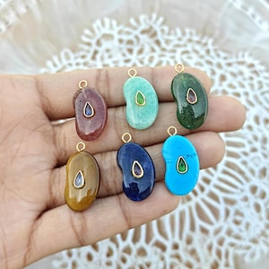 May include: A collection of seven gemstone pendants in various colors, including red, green, blue, and brown. Each pendant has a bean-like shape with a gold-colored bail and a contrasting gemstone inlay. The pendants are displayed against a soft, neutral background.