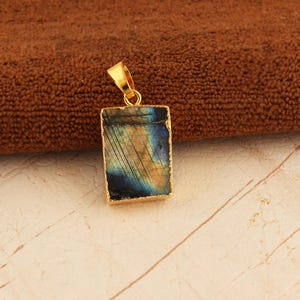 Può includere: Un ciondolo rettangolare in labradorite con castone e anello dorati. La pietra mostra colori iridescenti blu, verdi e oro. Il ciondolo è posto su uno sfondo marrone e crema.