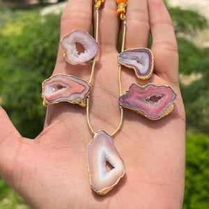 May include: A necklace with agate pendants in pink, white, and gold. The agate stones have a natural, crystal-like appearance and are set in gold-coloured metal. The necklace is displayed on a hand against a blurred green background.