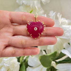 Puede incluir: Colgante en forma de corazón con una piedra preciosa de color rojo intenso, acentuado con metal dorado. El colgante presenta una pequeña piedra redonda y transparente en el centro y está suspendido de una fianza dorada. Los bordes del corazón están adornados con pequeñas cuentas doradas.