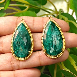 Puede incluir: Dos colgantes en forma de pera con piedras preciosas de color verde oscuro facetadas, engastadas en marcos de metal dorado con un borde de cuentas. Cada colgante incluye un pequeño lazo circular en la parte superior para sujeción.