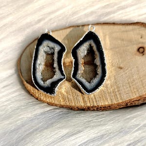 May include: Two matching geode pendants with a silver-colored trim. The pendants have a black and white outer layer with a crystal-filled center. The pendants are displayed on a wooden slice.
