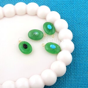May include: Four green oval-shaped gemstone pendants with gold-colored loops. Each pendant has a different center: a black oval, a turquoise oval, a blue oval, and a clear, iridescent center. The pendants are displayed on a white, wavy surface.