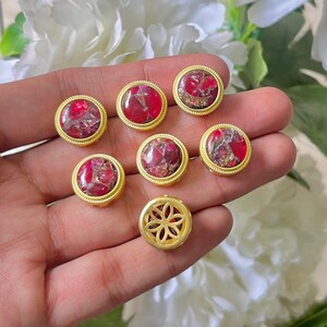 May include: A collection of gold-colored jewelry components. The components feature a round, red and gold stone set within a gold bezel. One component has a floral design. The components are displayed against a soft background.