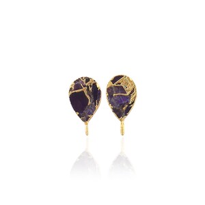 May include: A pair of teardrop-shaped earrings featuring deep purple amethyst stone slices with jagged, natural edges. Each stone is encased in a textured gold-tone metal frame with a small loop at the bottom for attaching additional jewelry components.