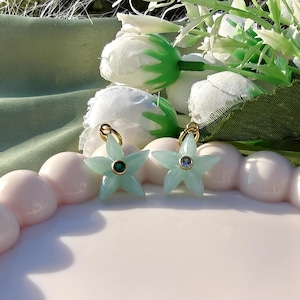 May include: Two light blue star-shaped earrings with gold hoops. Each earring has a central gemstone, one green and one blue. The earrings are presented on a pink, scalloped surface, with white flowers in the background.