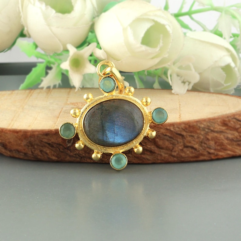 May include: A gold-colored pendant with an oval, iridescent, gray-blue stone at its center. The pendant is adorned with small, round, light-green stones and gold-colored accents. The pendant is displayed on a wooden surface with white flowers in the background.