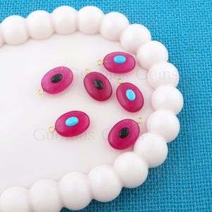 May include: Six oval-shaped gemstone beads in shades of magenta, black, and turquoise. Each bead has a gold-colored loop at the top for stringing. The beads are displayed on a white, wavy-edged tray against a blue background.