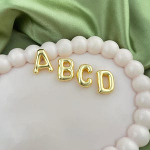 Może przedstawiać: Złote metalowe litery piszące ABCD. Litery są wyświetlane na jasnoróżowej, okrągłej powierzchni z białym, koralikowym obramowaniem. Tło to zielona, jedwabista tkanina.