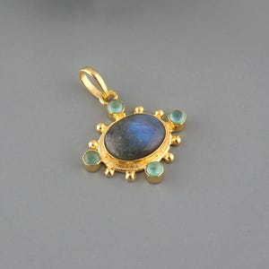 May include: A gold-colored pendant featuring a central oval-shaped, iridescent blue-gray stone, surrounded by smaller, round, light green stones. The pendant has a decorative, sunburst-like design and a loop for a chain.