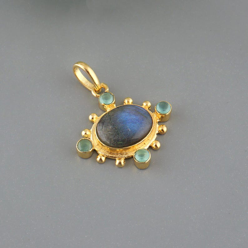 May include: A gold-colored pendant featuring a central oval-shaped, iridescent blue-gray stone, surrounded by smaller, round, light green stones. The pendant has a decorative, sunburst-like design and a loop for a chain.