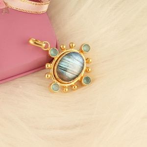 May include: A gold-colored pendant with an oval, iridescent stone at its center, surrounded by small, light blue gemstones and gold accents. The pendant is on a white, fluffy surface, next to a pink gift box with a gold ribbon.