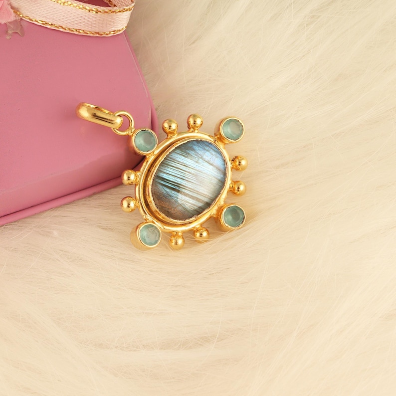 May include: A gold-colored pendant with an oval, iridescent stone at its center, surrounded by small, light blue gemstones and gold accents. The pendant is on a white, fluffy surface, next to a pink gift box with a gold ribbon.