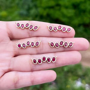 May include: Close-up of several gold-coloured jewellery pieces, each with a row of oval, deep red gemstones. The gemstones are set in a gold metal frame. The jewellery is displayed against a blurred green background.