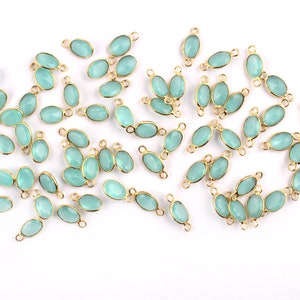 Può includere: Una collezione di connettori ovali di gemme blu chiaro con montature in metallo dorato. I connettori sono sparsi su una superficie bianca, mettendo in risalto il loro design delicato. Questi componenti di gioielleria sono ideali per creare collane, bracciali e orecchini.
