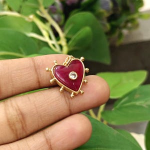 May include: A gold-colored heart-shaped pendant with a deep red center and a small, clear gemstone. The pendant is surrounded by gold accents and small, round embellishments. The pendant is held in a hand against a backdrop of green leaves.
