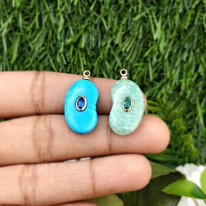 May include: Two gemstone pendants. One is turquoise blue with a blue and gold accent, the other is light green with a green and gold accent. Both have a gold loop at the top for hanging.
