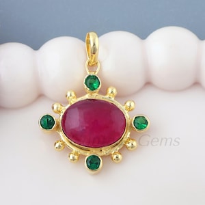 May include: A gold-colored pendant with a large, oval, deep red gemstone at its center. The pendant is adorned with smaller green gemstones and gold-colored accents. The design is symmetrical and features a loop for a chain.