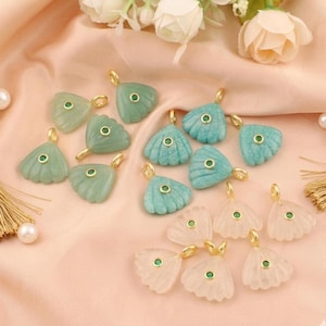 May include: A collection of gemstone pendants in various colors, including light green, turquoise, and translucent pink. Each pendant features a gold-colored bail and a small, round, green gemstone. The pendants are arranged on a pink fabric.