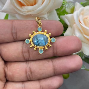 May include: A gold pendant with an oval, blue-grey stone at the center, surrounded by small, light blue stones and gold accents. The pendant is held up by a gold loop. The design is ornate and detailed, suitable for jewelry.